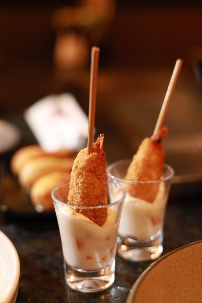 Dois copinhos de vidro com molho branco cremoso, cada um com um camarão empanado espetado num palito de bambu