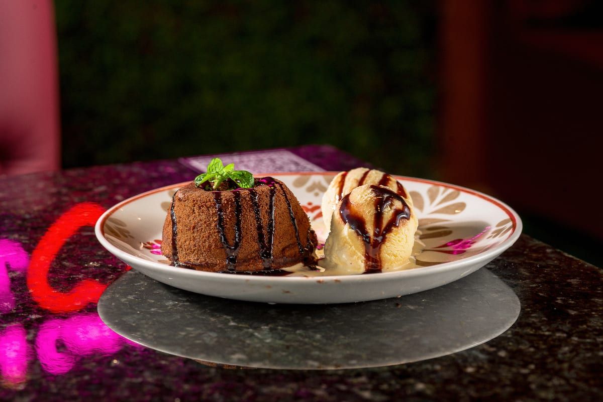 Bolinho de chocolate com calda escura ao lado de duas bolas de sorvete de creme, em prato floral