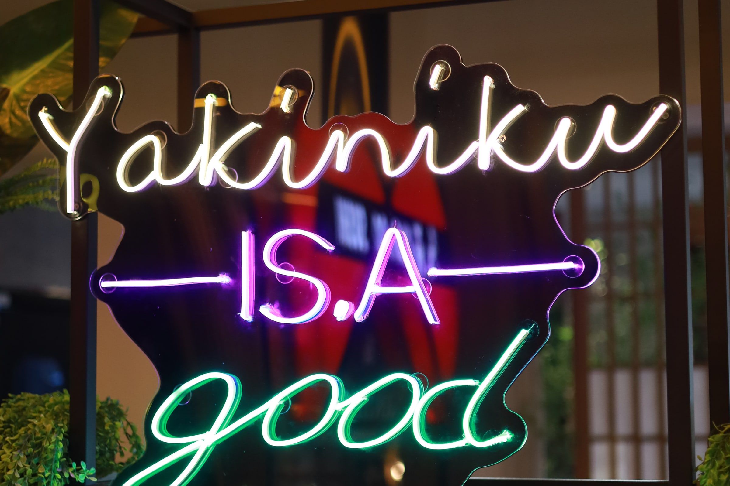 Letreiro de neon escrito Yakiniku is a good idea em roxo e verde, entre as folhagens da parede do salão