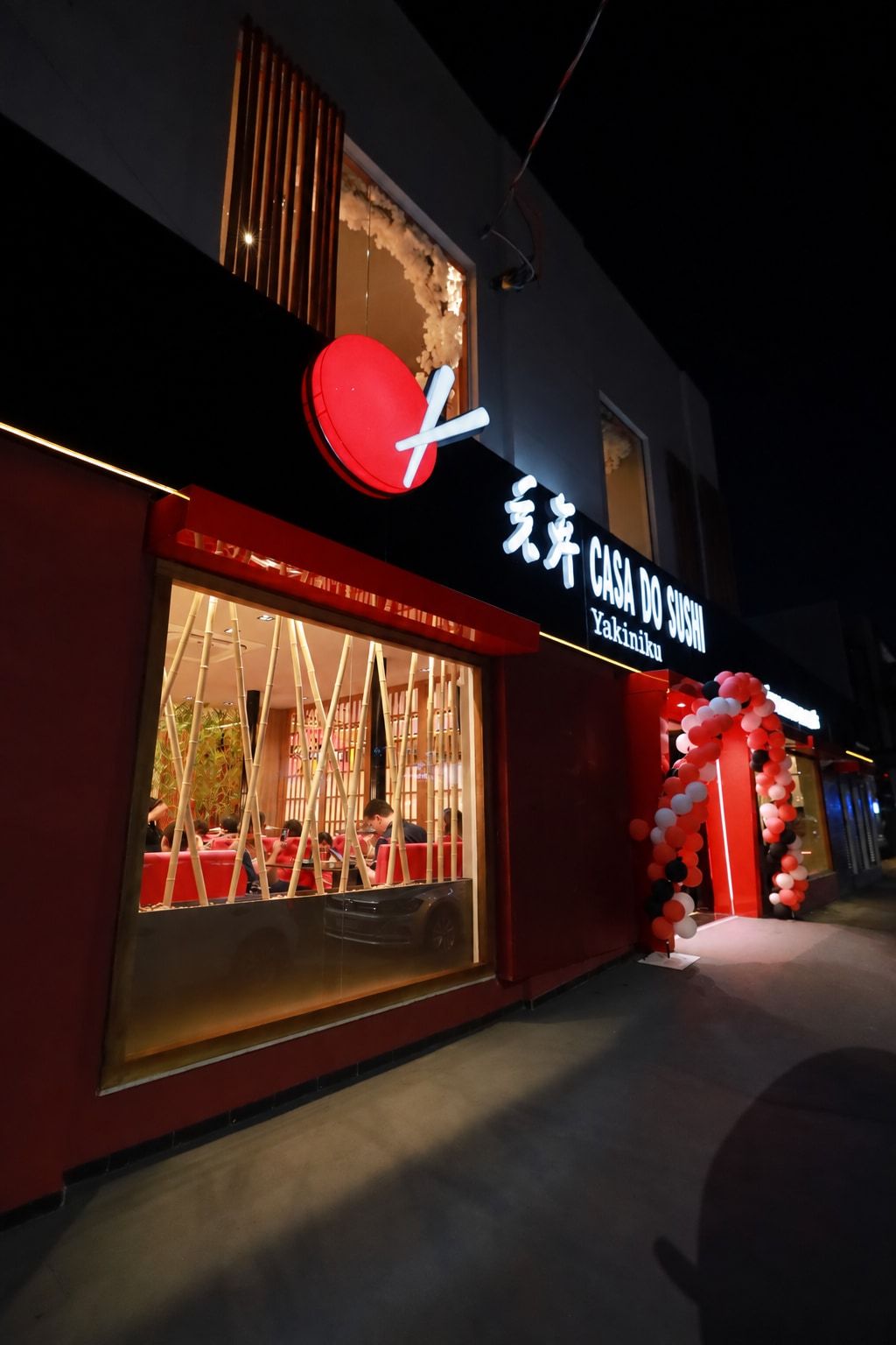 Fachada à noite vista da calçada, com o logo vermelho aceso, a vitrine de bambus e o arco de balões na entrada