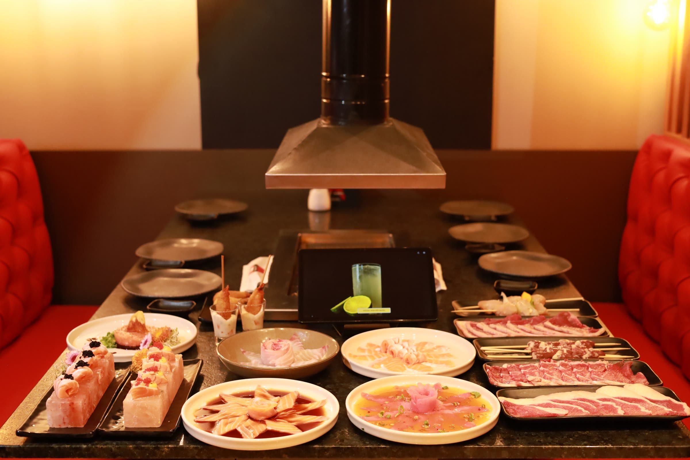 Mesa de rodízio posta sob a coifa, com bandejas de carne crua fatiada, pratos de sashimi e acompanhamentos, entre bancos capitonê vermelhos