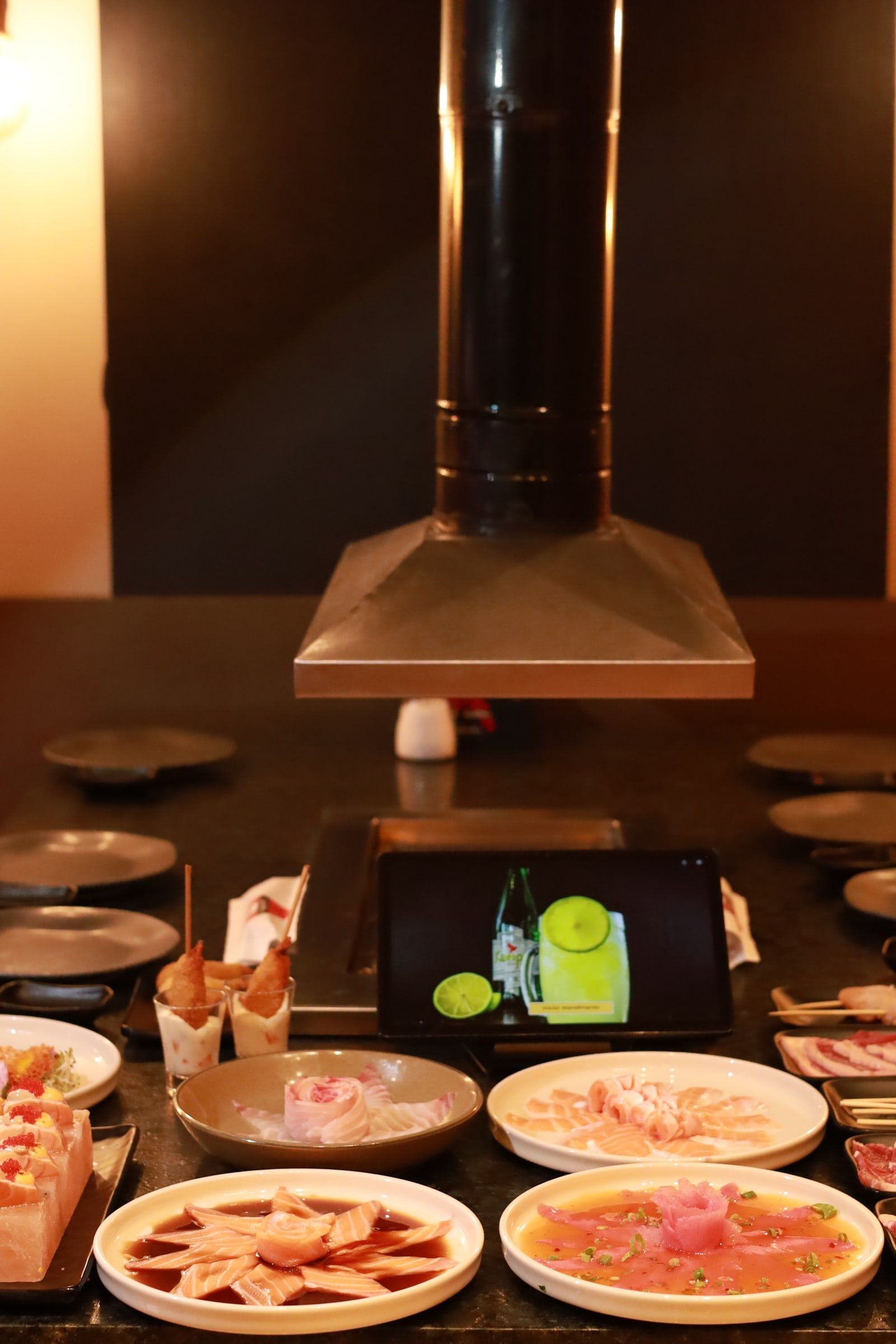 Coifa de cobre descendo sobre a mesa de granito posta com pratos de sashimi, cones de temaki e o tablet do pedido