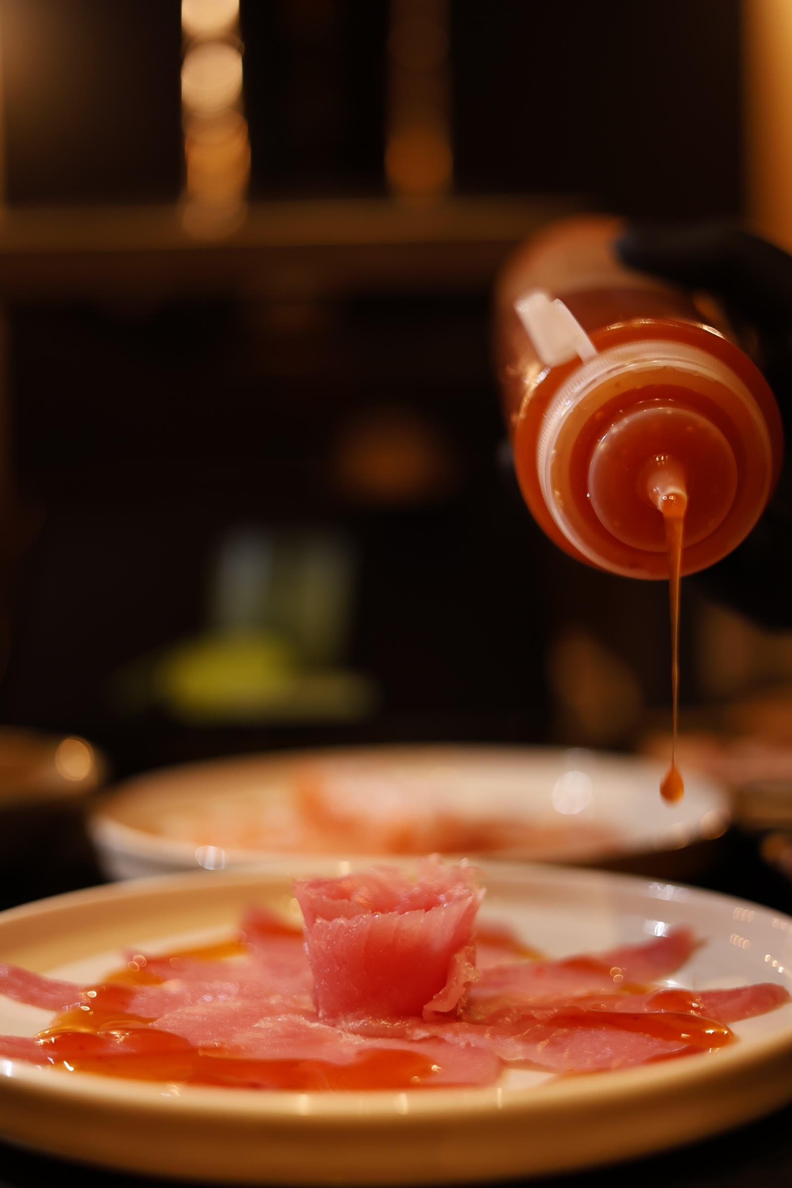Mão enluvada despeja um fio de molho alaranjado sobre o carpaccio de peixe com rosa ao centro