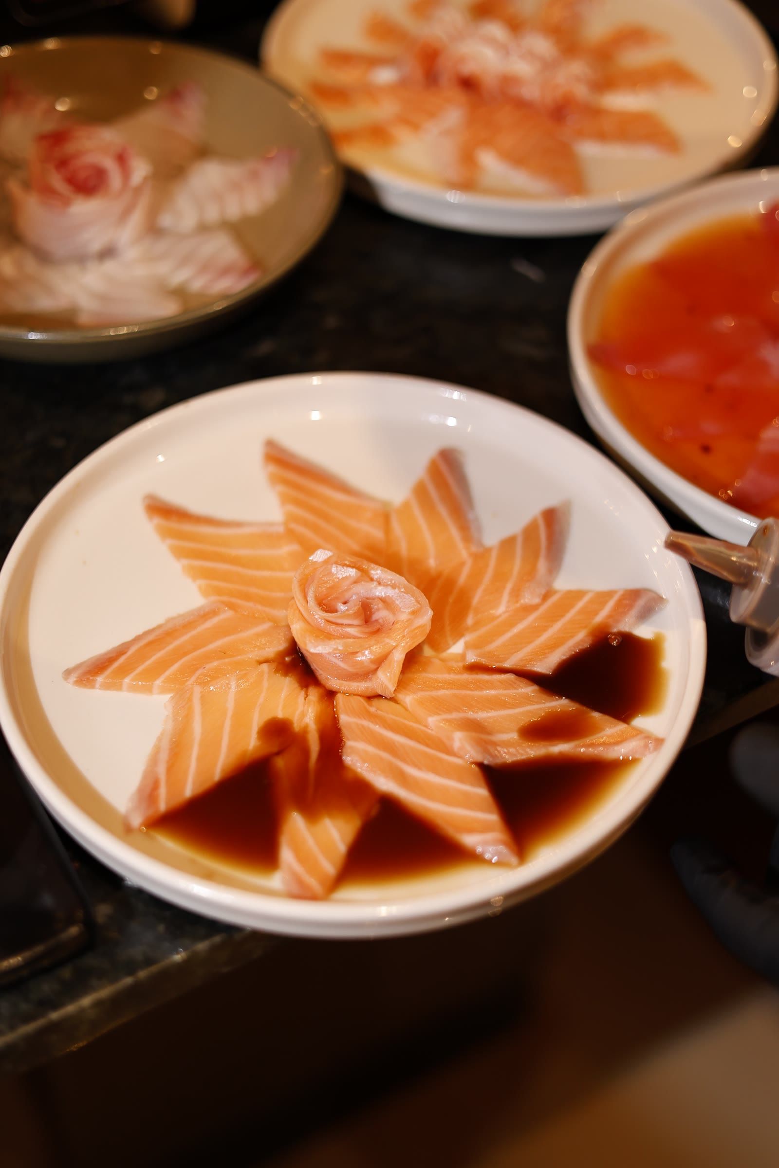 Prato de sashimi de salmão em flor sobre shoyu, cercado por outros pratos no balcão