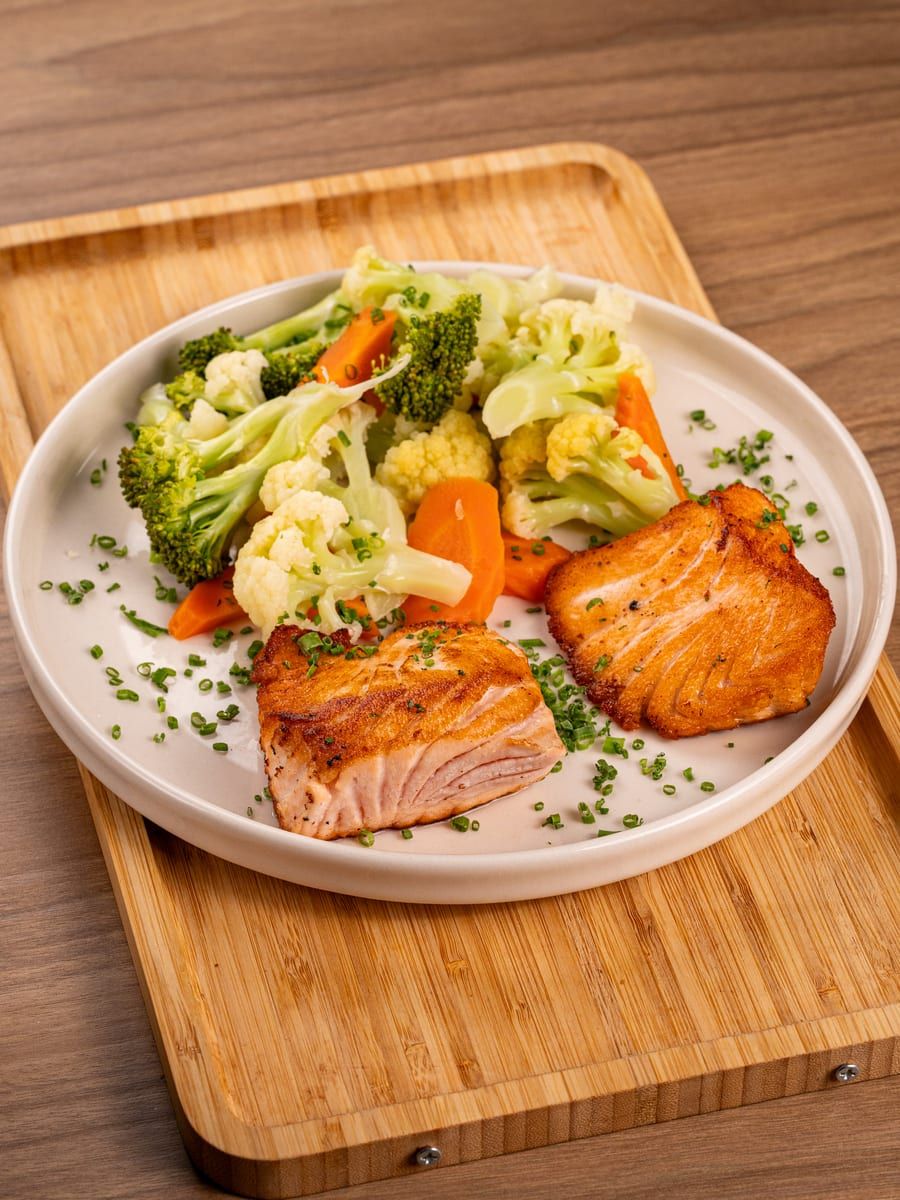Postas de salmão grelhado ao lado de legumes salteados, em prato branco sobre tábua de bambu