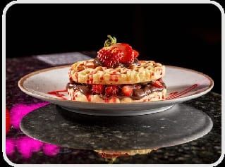 Waffle em camadas com calda e um morango inteiro no topo, em prato sobre mesa de pedra